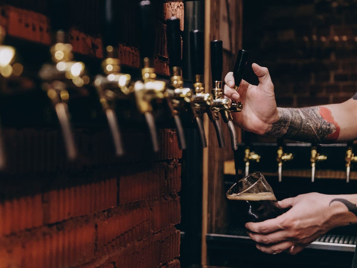 Hands pouring a dark beer from a row of gold-toned draft faucets on a brick wall